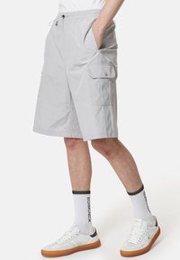 Pantaloni cargo grigio chiaro con un cinturino elastico in vita, tasche laterali e una vestibilità comoda. Indossati con scarpe da ginnastica bianche e calze con dettagli neri.