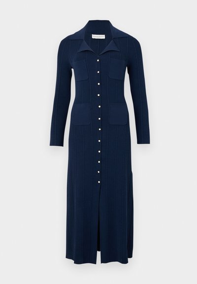 Abito lavorato a maglia blu navy con colletto, bottoni frontali e due tasche sul petto. Presenta una texture a costine verticali e uno spacco frontale per la libertà di movimento.