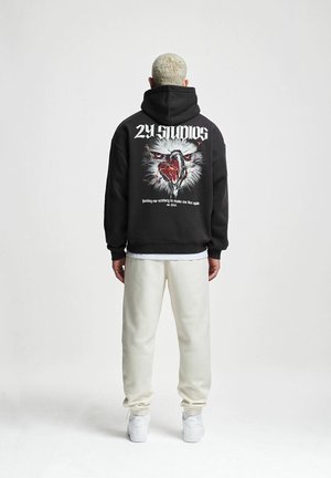 Black hooded sweatshirt with a graphic of an eagle and text on the back, paired with light beige sweatpants and white sneakers.