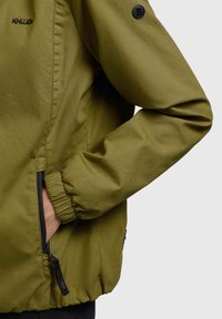 Olive green jacket with a lightweight, smooth texture. Features elastic cuffs, side zippers, and a logo on the shoulder.