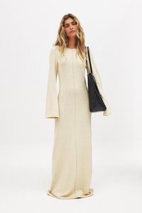 Robe tricotée longue couleur crème avec des manches évasées et une couture centrale. Associée à un sac à main noir pour le contraste. Texture lisse.