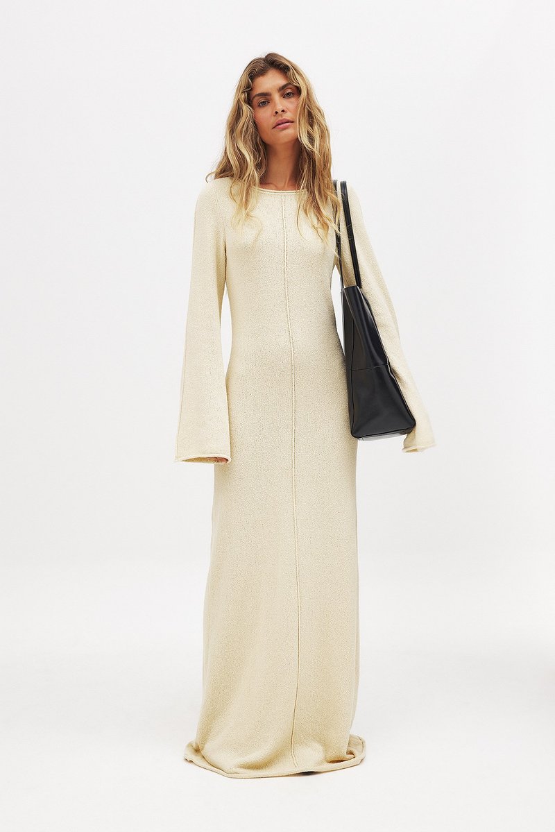 Robe tricotée longue couleur crème avec des manches évasées et une couture centrale. Associée à un sac à main noir pour le contraste. Texture lisse.