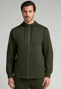 Sudadera con cremallera de color verde oscuro, con capucha, hecha de un tejido suave. Cuenta con bolsillos laterales, puños elásticos y cordones ajustables en la capucha.