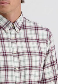 Lichte, knoopsluiting blouse in het wit met een bordeauxrood en teal geruit patroon; voorzien van een puntige kraag en een borstzak aan de linkerkant.