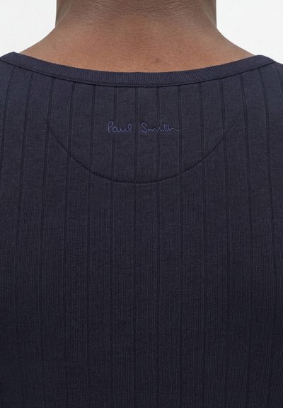 Κοντινό πλάνο στο πάνω μέρος της πλάτης ενός ατόμου που φοράει σκουρόχρωμο ριμπ ύφασμα με το «Paul Smith» κεντημένο με μικρά, διακριτικά γράμματα κοντά στη λαιμόκοψη.