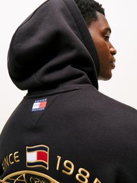 Sudadera negra con capucha, que presenta un parche con el logo en la parte superior de la espalda y bordados en oro, incluyendo el año 1984 y un diseño de bandera en bloques de color.
