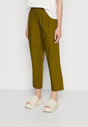 Persoon die olijfgroene cropped broek draagt met een elastische tailleband en witte platformsandalen tegen een effen achtergrond.