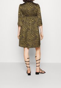 Khaki groene jurk met een zwart zebra-streep patroon, halflange mouwen, getailleerde taille en een gestructureerde stof. Gekleed met zwarte bandjesandalen.