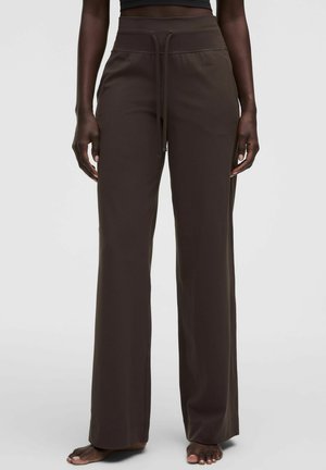 Persona que lleva pantalones anchos de pierna en color marrón oscuro con cintura ajustable y una camiseta negra corta, de pie descalza sobre un fondo liso.