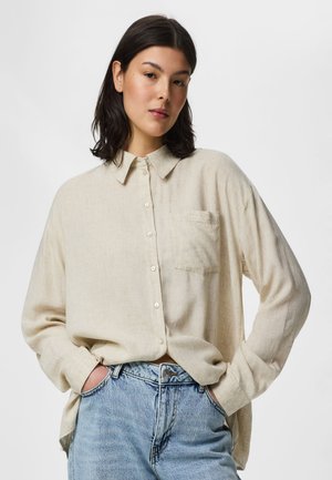 Jonge vrouw met donker haar, dragend een beige blouse met knopen en lichtblauwe spijkerbroek, handen ontspannen in de zakken, tegen een effen achtergrond.