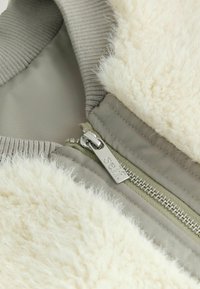 Kremowa kurtka z sztucznego futra z jasnoszarym podbiciem. Posiada srebrny zamek błyskawiczny i prążkowany kołnierz. Miękka faktura i dopasowany krój.