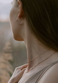 Roségoldene Creolen, geschichtete zarte Ketten auf einem gebräunten Hals, glattes Haar teilweise zusammengebunden, sanftes Licht hebt die Texturen hervor.