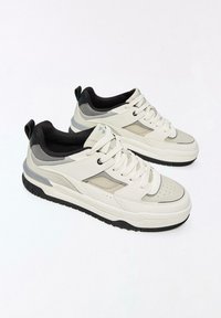 Fehér sportcipő szürke hálós és díszítő elemekkel. Texturált gumitalppal, fűzős elejű és párnázott szegéllyel a kényelemért.