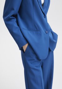 Traje azul a medida que presenta un blazer estructurado con dos botones y bolsillos laterales, combinado con pantalones a juego, textura de tela suave.