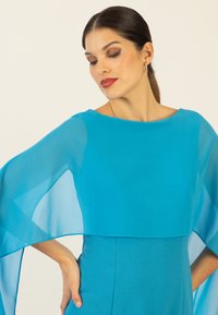 Ein langärmliges Kleid aus leuchtend blauen Stoff, mit einem fließenden, durchsichtigen Überzug, einem runden Ausschnitt und einer glatten, figurbetonten Silhouette.