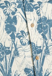 Blauwe bloemenprint op een beige stoffen shirt, met houten knopen en een gestructureerde afwerking. Close-up van de ontwerpelementen en de plaatsing van de knopen.