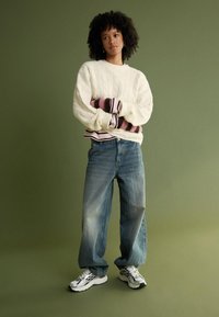 Krämfärgad kabelstickad tröja över en randig rosa och vit topp, i kombination med lösa blå jeans och silverfärgade sneakers. Grön bakgrund.