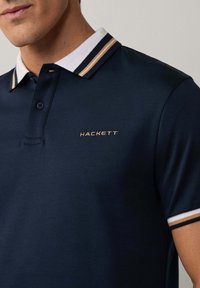 Námořnická polo košile s bílým límcem, obsahující zlaté a námořnické pruhy, vyšitým logem "HACKETT" ve zlaté barvě na levé části hrudi, krátké rukávy.