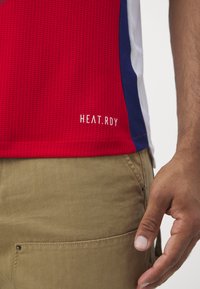T-shirt de sport rouge avec une texture en maille, présentant des accents latéraux bleus et une étiquette blanche "HEAT.RDY" en bas. Un pantalon kaki est visible.