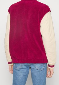 Fleecetröja med en klar magentaröd rygg och krämfärgade ärmar. Har ribbade muddar med vinröda detaljer. Mjuk textur och avslappnad passform.