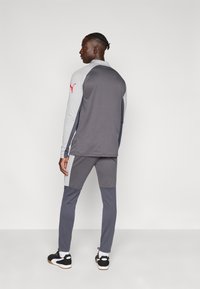 Γκρι αθλητική φόρμα με αντίθετα ανοιχτογκρι πάνελ, εφαρμοστό σχέδιο και κόκκινο λογότυπο στο μανίκι. Μαύρα και λευκά αθλητικά παπούτσια.