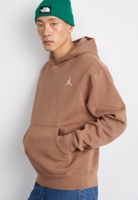 Sweat à capuche brun avec poche kangourou et logo blanc. Porté avec un jean bleu et un bonnet en tricot vert. Tissu lisse et coupe décontractée.