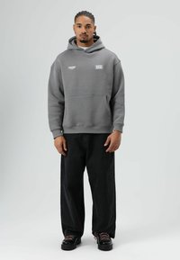 Grijze hoodie met kap en voorzak, voorzien van witte merkemblemen. Gecombineerd met zwarte wijde jeans en zwarte schoenen met roze veters.