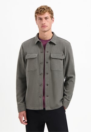 Grijze button-up jas met twee borstzakken, textuurstof en een kraag. Gedragen over een paarse shirt, met een recht gesneden ontwerp.