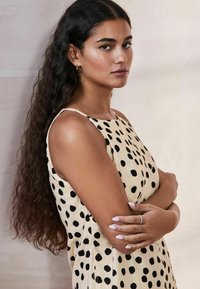 Soevaba beige kleit mustade täppidega, tekstureeritud kangas. Mudel, kellel on pikad lainelised juuksed, käed ristis, kannab sõrmuseid sõrmedes.