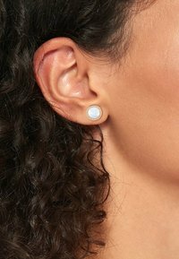 Boucles d'oreilles rondes avec une incrustation bleu clair et brillante, encadrées par un cercle métallique doré. Le design est mis en valeur par une coiffure foncée et bouclée texturée.
