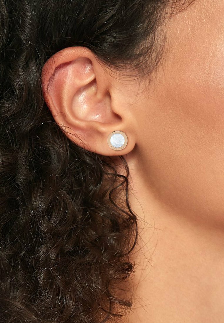 Boucles d'oreilles rondes avec une incrustation bleu clair et brillante, encadrées par un cercle métallique doré. Le design est mis en valeur par une coiffure foncée et bouclée texturée.