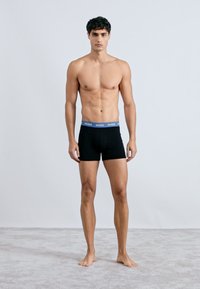 Boxer shorts neri con cintura a logo blu su un modello maschile senza camicia. Tessuto morbido, forma aderente, in piedi su un tappeto chiaro.
