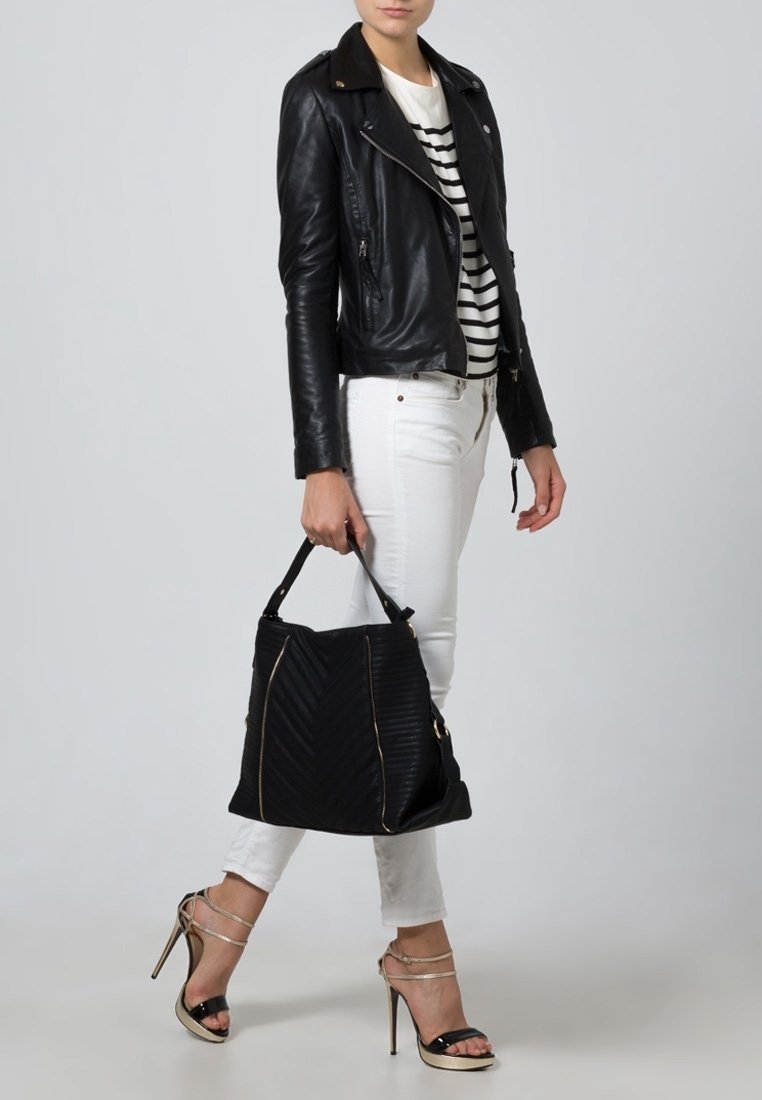 Femme portant une veste en cuir noire, un haut rayé, un pantalon blanc et des sandales à talons beiges, tenant un sac à main noir à ses côtés.