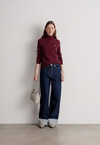 Burgunderfarbener Rollkragenpullover kombiniert mit weit geschnittenen, dunklen Jeans mit Saum. Das Modell hält eine beige Tasche und steht vor einem neutralen Hintergrund.