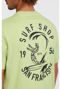 Camiseta de algodón verde claro con un gráfico negro de un surfista y una ola, con el texto "SURF SHOP" y "SAN FRANCISCO" en letras negritas.