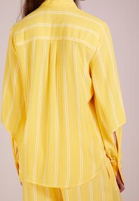 Maglietta a righe gialle con linee bianche, vestibilità ampia, colletto e maniche aperte. Tessuto morbido, dettaglio della piega sul retro, progettata per un look rilassato.