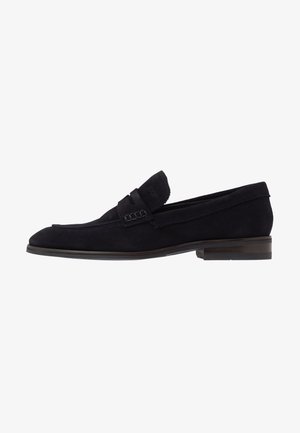 Mocasín de ante negro para hombre con un tacón bajo y una correa decorativa en la parte superior, mostrado de perfil contra un fondo blanco.
