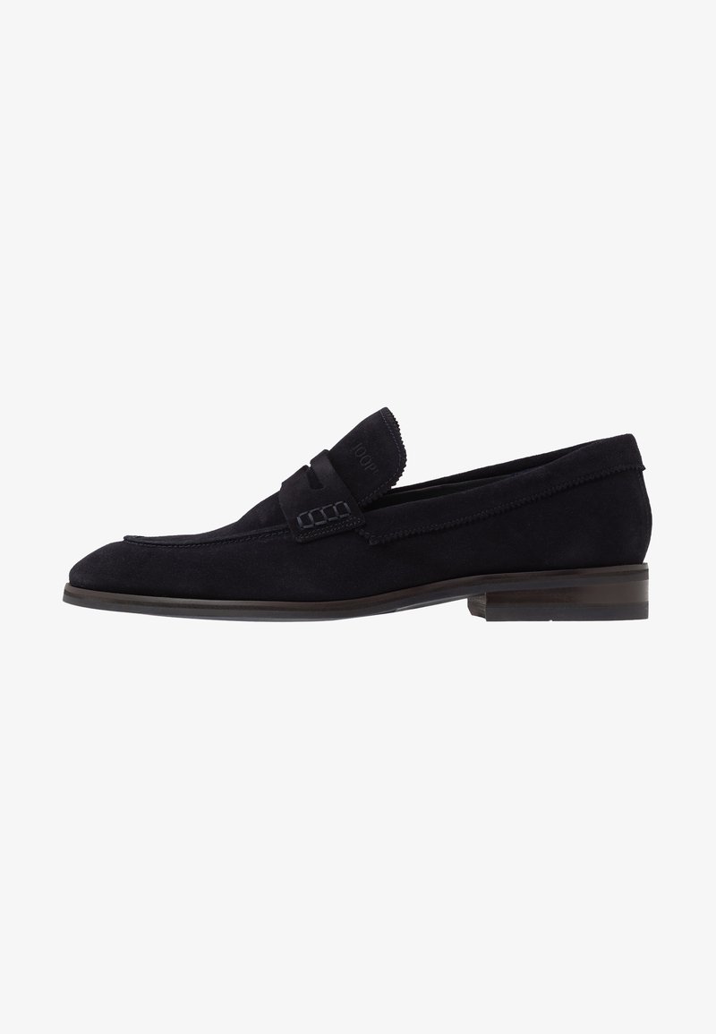 Mocassin pour homme en daim noir avec un petit talon et une lanière décorative sur le dessus, présenté en profil latéral sur un fond blanc.