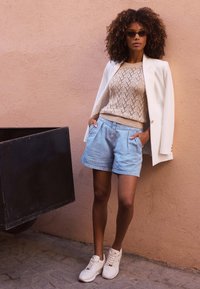 Vit kavaj över en beige stickad tröja med diamantmönster, parat med ljusblå denimshorts och vita sneakers.