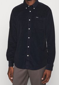 Chemise boutonnée en velours côtelé marine à manches longues, avec une poche poitrine simple, des boutons blancs et une petite étiquette de marque sur la poche.