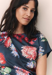 Blouse fleurie en satin bleu marine à manches courtes. Présente de grands motifs floraux rouges et bleus. Texture lisse avec une coupe décontractée.