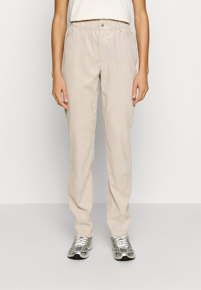 Personne portant un pantalon beige à taille élastique et des baskets argentées, debout devant un fond clair uni.