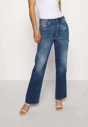 Kobieta ubrana w wyblakłe niebieskie dżinsy o prostych nogawkach, biały top na ramiączkach oraz białe sandały na obcasie z odkrytymi palcami, stojąca na jednolitym tle.