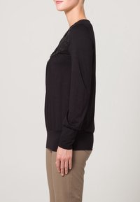 Svart långärmad topp tillverkad av mjukt material, med en texturerad spetsdetalj och figurnära silhuett. S änkt axelsöm och ribbad nederkant.