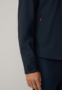 Giacca softshell blu navy con tasca con cerniera, polsini regolabili e texture liscia. Accento rosso visibile sulla cerniera della tasca.