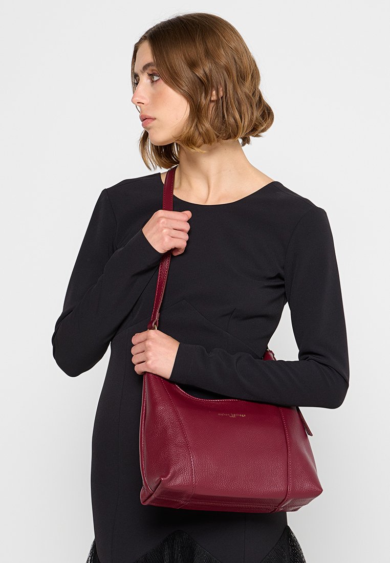 Sac à épaule en cuir bordeaux avec une texture douce, une forme légèrement structurée et une longue bandoulière ; présente un branding subtil.