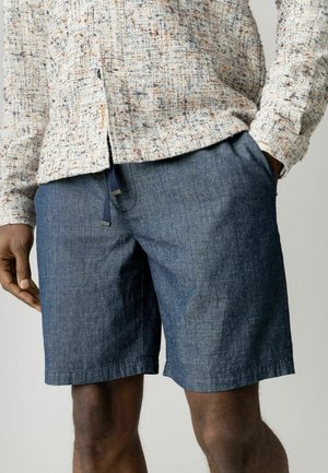 Férfi visel texturált krémszínű, gombolós ingben és kék farmer rövidnadrágban, kezét zsebében tartva, egyszínű háttér előtt.