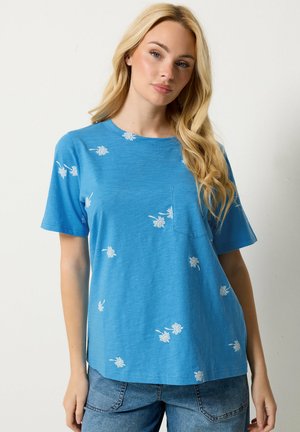 Mujer rubia con camisa de manga corta azul con pequeños estampados de flores blancas y vaqueros azules sobre un fondo liso.