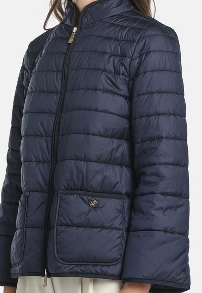 Donna che indossa una giacca trapuntata blu navy con grandi tasche frontali e chiusura con zip dorata, spalle leggermente cadenti.