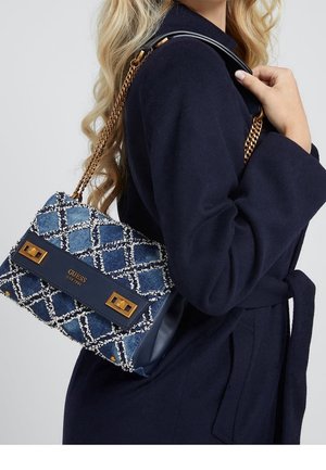 Vrouw met marineblauw jas houdt een blauw-wit patroon Guess handtas met gouden kettingriem over haar schouder.